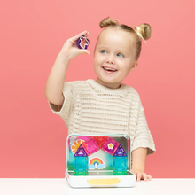 Load image into Gallery viewer, A young child with blonde pigtails smiles while holding a colorful magnetic building block piece above an open case of magnetic tiles on a white table, set against a solid pink background.