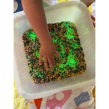 Load image into Gallery viewer, A child's hand touching glowing green sand in a clear plastic tub, with multicolored beads and glitter inside.