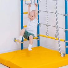 Load image into Gallery viewer, A baby wearing a white t-shirt and green shorts hangs from a blue and yellow indoor playground structure with gray rings and ropes, sitting on a yellow foam mat.