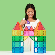Load image into Gallery viewer, A young girl with long dark hair, wearing a white top, smiles as she builds a colorful translucent castle with magnetic building blocks on a white table against a solid light green background.