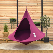 Load image into Gallery viewer, A hanging teardrop-style chair in a rich magenta-purple color, suspended by a metal chain in front of a light wood paneled wall. The chair features a small white tag on the side and is set against a background of potted plants and a wooden floor.