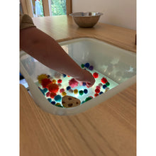 Load image into Gallery viewer, A child's hand reaching into a clear plastic sensory tub filled with colorful pom-poms and a small cookie-shaped toy, set on a light wood table.