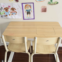 Load image into Gallery viewer, A light wood children's table with two matching wooden chairs in a classroom or playroom setting, with children's artwork hanging on the wall behind it.