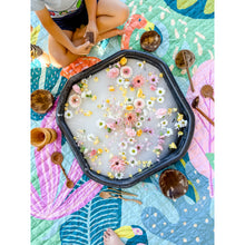 Load image into Gallery viewer, A top-down view of a child sitting on a colorful quilted blanket next to a black hexagonal tub filled with water, flowers, and floating petals. Wooden spoons, coconut shells, and other natural items are arranged around the tub on a bright patterned blanket.
