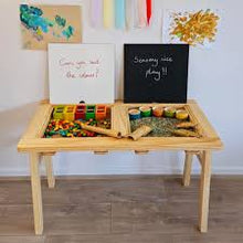 Load image into Gallery viewer, Children's play area with wooden toys on a table, framed pictures on the wall, and a blackboard with text.
