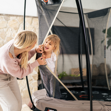Load image into Gallery viewer, A woman and a young girl smiling and jumping on a trampoline inside a safety enclosure net in a living room setting.