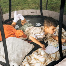 Load image into Gallery viewer, A young boy lies on a trampoline, reading a book while surrounded by cozy blankets, pillows, and a plush toy. He is wearing a beige t-shirt with white stripes, green shorts, and white socks.