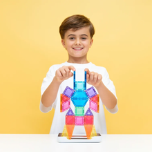 Load image into Gallery viewer, A smiling young boy in a white t-shirt stands behind a colorful magnetic tile structure built on a tablet, against a solid yellow background.
