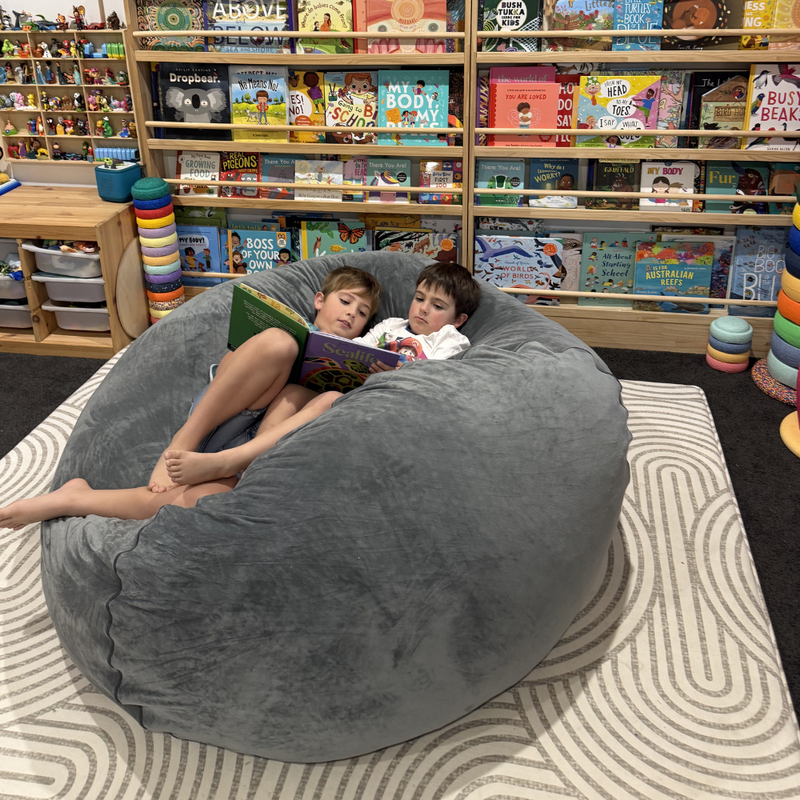 Two children sitting in a large gray bean bag reading books in a bookstore.