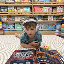Load image into Gallery viewer, Child wearing headphones lying on a rug in a bookstore, reading a book.
