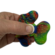 Load image into Gallery viewer, A hand holding a three-spoke silicone fidget spinner with a multicolor rainbow pattern, featuring raised textured bumps on the blades and a smooth white center hub.
