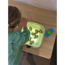 Load image into Gallery viewer, A child in a mint green dress leans over a glowing light box containing several small eggs, with a few blue and green pieces on the table above.
