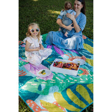 Load image into Gallery viewer, A woman in a blue dress holds a baby while sitting on a colorful outdoor picnic blanket on grass, with a young girl in a white dress sitting beside them and a box of fruit in front.
