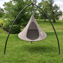 Load image into Gallery viewer, A hanging tree swing hammock with a beige canopy and dark gray mesh window, suspended from a black metal arch frame on a green grass lawn with trees in the background.
