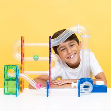 Load image into Gallery viewer, A smiling young boy with dark hair wearing a white t-shirt leans behind a colorful magnetic marble run toy set on a white table against a solid yellow background.
