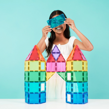 Load image into Gallery viewer, A young girl with long dark hair, wearing a white sleeveless top, smiles while holding a pair of translucent blue magnetic glasses in front of her eyes. In front of her, she has built a colorful castle structure using translucent rainbow-colored magnetic building blocks against a solid light blue background.
