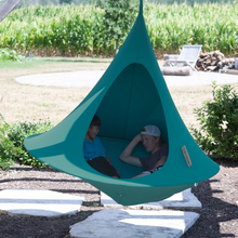 Load image into Gallery viewer, A teal outdoor hanging hammock pod with two people sitting inside, photographed in a backyard setting with a cornfield and patio visible in the background.
