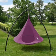 Load image into Gallery viewer, A purple hanging tree swing hammock suspended from a black metal arch frame, set up on a green grass lawn with trees in the background.
