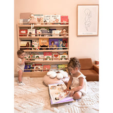 Load image into Gallery viewer, Two children in a room with a bookshelf filled with books and a sofa.
