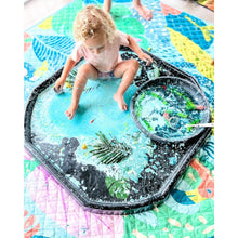 Load image into Gallery viewer, A toddler sits inside a black octagonal sensory play tray filled with blue water and toy pieces, placed on a colorful quilted blanket. A matching round tray with more water and green leaves sits beside them.
