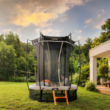 Load image into Gallery viewer, A large round trampoline with an attached black safety enclosure net and jump mat, set in a backyard on green grass. A child is visible inside the enclosure, and an orange step ladder stands in front.
