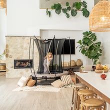 Load image into Gallery viewer, A woman stands inside a Vufu oval trampoline with a black safety net in a bright, modern living room with a stone fireplace, wooden floors, and a dining table.
